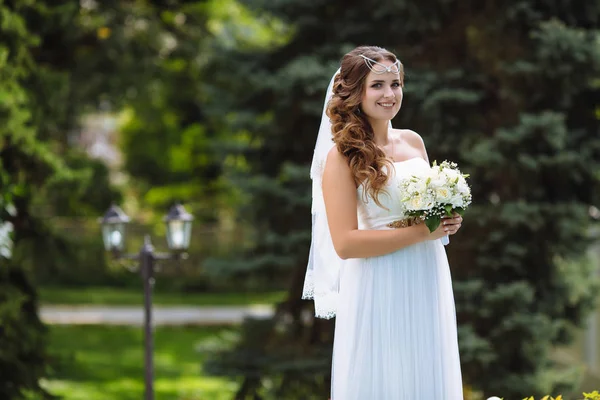 Retrato de uma jovem noiva em um longo vestido de noiva branco fluindo e véu, fica no parque contra as árvores, segurando um buquê de rosas em suas mãos . — Fotografia de Stock