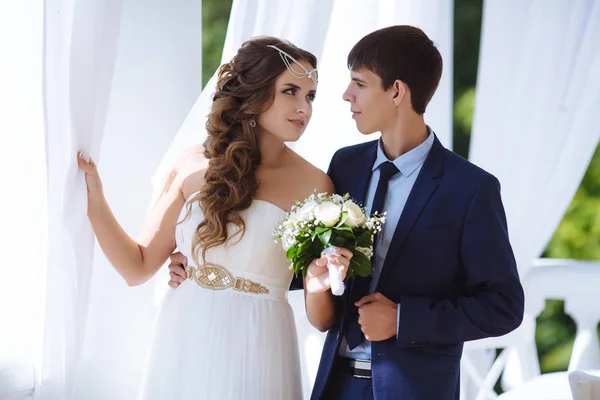 Die schöne Braut trägt ein Kleid im griechischen Stil, ihr Haar ist mit Perlen verziert, sie hält einen klassischen Hochzeitsstrauß weißer Rosen in der Hand und blickt ihren schlanken, großen Mann in einem blauen Geschäft an — Stockfoto