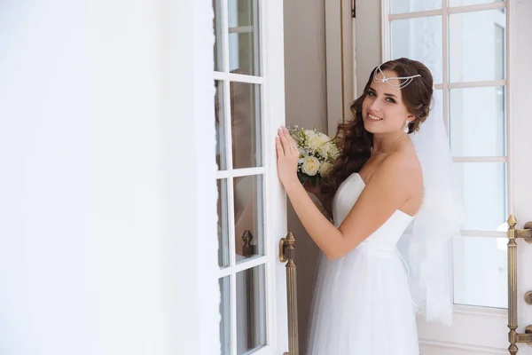 Uma noiva doce na imagem de uma deusa grega decorou seu cabelo com uma lapela, pegou um véu de renda e um vestido de casamento leve sorri e posa perto da porta de vidro onde seu reflexo é visível . — Fotografia de Stock