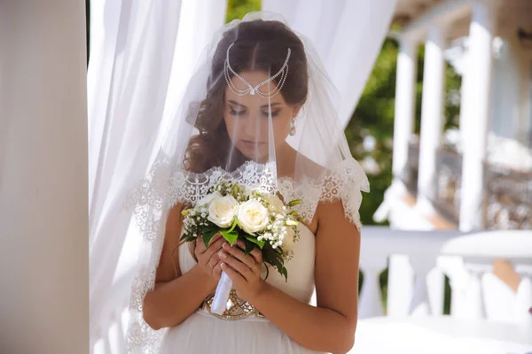 Egy gyönyörű lány viselt egy esküvői ruha és egy csipke fátyol, amelyekre ő a fej és arc, gondosan kezében egy esküvői csokor Rózsa mindkét kezével, megcsodálta őket. — Stock Fotó