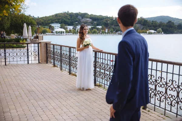 Vacker flicka i bröllop klänning leenden att hennes brudgum. Paret promenader vid floden, beundrar staden från den andra stranden, jublar på deras bröllop och en varm dag. — Stockfoto
