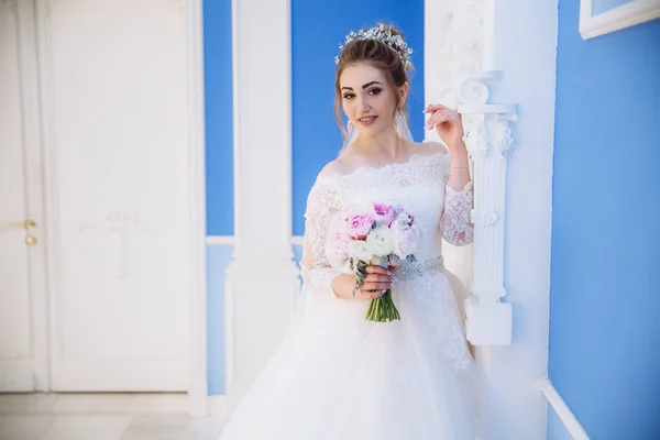 Frumoasa blonda pozand langa o coloana alba intr-o rochie de mireasa cu un buchet de bujori roz . — Fotografie, imagine de stoc