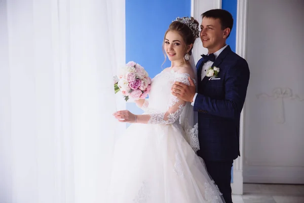 A girl in a wedding dress and with a bouquet of peonies pushes the curtain on the window. Behind her, a man in a blue suit is hugging her. — Stock Photo, Image