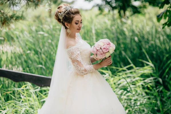 A menina está andando em um vestido de renda exuberante em um campo elegante com grama alta no dia do casamento. Um belo bando de espiões na mão das noivas . — Fotografia de Stock