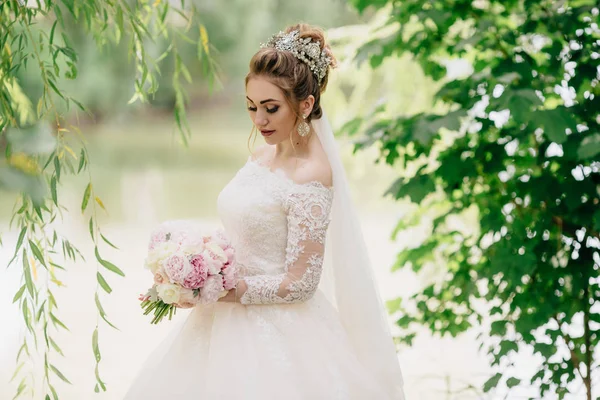 Porträt einer Braut, die in der Natur wandelt. , blickt das Mädchen nach unten und zeigt ihr wunderschönes weiches Make-up und lange Wimpern. eine Braut mit einem Strauß in der Hand geht zwischen Bäumen. — Stockfoto