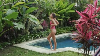 İnce güzel kız tropikal yeşil arıyorsunuz havuz kenarında duruyor ve kırmızı bitkiler ile açık bir arka renk parlak bir mayo giyiyor, kısa kıvırcık saçlı, ince uzun bacakları ve yuvarlak eşek vardır.