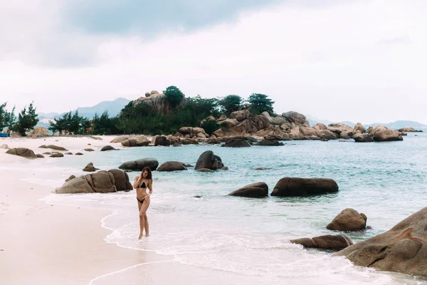 Ein schönes Mädchen spaziert an einem einsamen Strand zwischen großen Felsbrocken und viel Grün entlang. es berührt mit seinen schlanken Beinen blau transparentes warmes Wasser. — Stockfoto