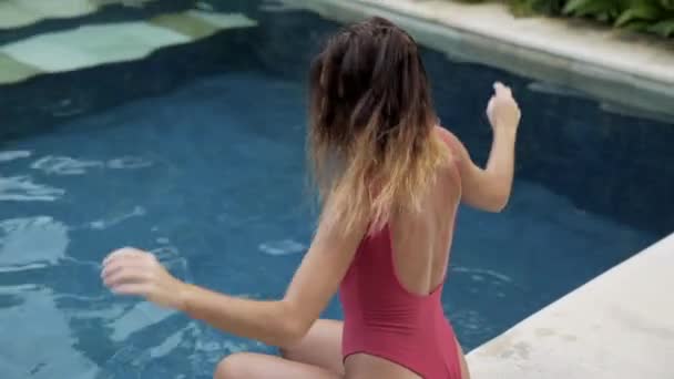View from the back of a beautiful young tanned girl with short hair in an open swimsuit sits at the edge of the pool, has a round sexy big ass, and sports back, slow-motion shooting, close-up. — Stock Video