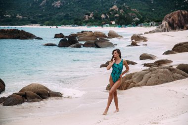 İnce uzun boylu güzel model siyah saçlı yeşil yüksek dağlar zemin ve kaya parçaları karşı masmavi plaj ve kıyı sahilde, manzaranın yürür.