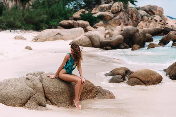 Schlanke attraktive junge Mädchen ruht auf einem weißen Sandstrand, sitzt auf einem Stein vom Wasser umgedreht, vor dem Hintergrund der großen Fragmente von Felsen. — Stockfoto