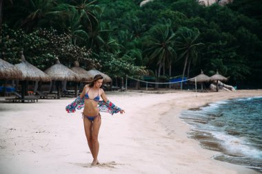 İnce, bronzlaşmış bir genç uzun boylu kız mayoyla beyaz kumlu bir plaj üzerinde yürür, düzgünce bakiyesinin bir parlak bikini mayo ve renkli bir taraf için ellerini koyarak ona çorap üzerinde yürüyor
