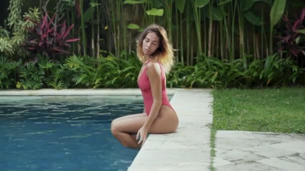 Das Model mit dem runden großen sexy Arsch sitzt am Pool und beobachtet die Wassertropfen, die aus ihren Händen abfließen, trägt ein modernes Stück hellen Badeknochen, der Wind spielt mit ihrer kurzen — Stockvideo