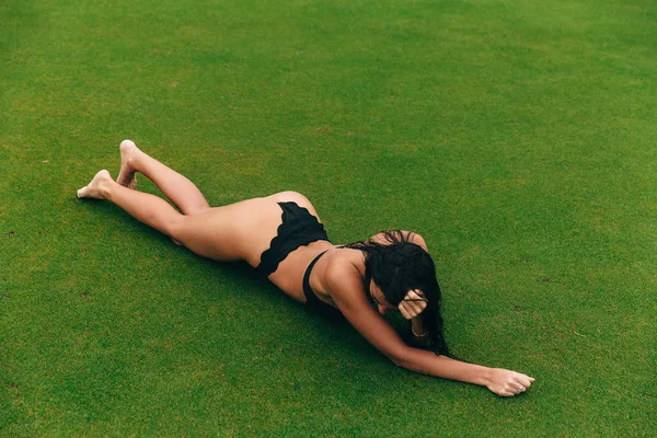 Vista dall'alto di una ragazza sottile e abbronzata con i capelli scuri si trova in un costume da bagno nero sull'erba verde, il suo viso non è visibile . — Foto Stock