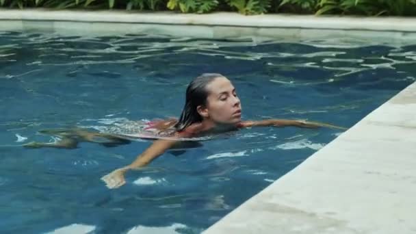 Uma linda morena com cabelo molhado curto em um maiô de framboesa salta da piscina azul, entra em seus braços, tem um peito grande, uma cintura fina e uma bunda redonda, um tiro em câmera lenta . — Vídeo de Stock