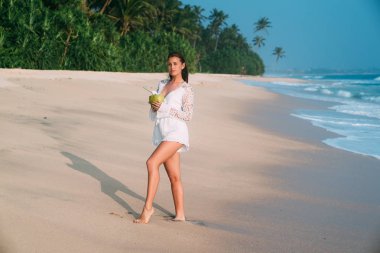 Güzel bir genç kız şık plaj kıyafetlerini giymiş pembe kumlu plaj boyunca yürüyor, taze Hindistan cevizi içecekler, tropikal orman ve deniz masmavi bir deniz hayran