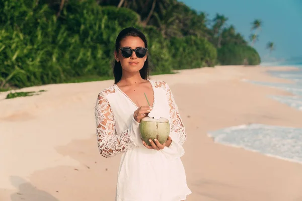 Hermosa joven bronceada brillante en la playa con una camisa blanca y gafas de sol de moda, sosteniendo un coco fresco con un tubo, caminando a lo largo del océano . — Foto de Stock