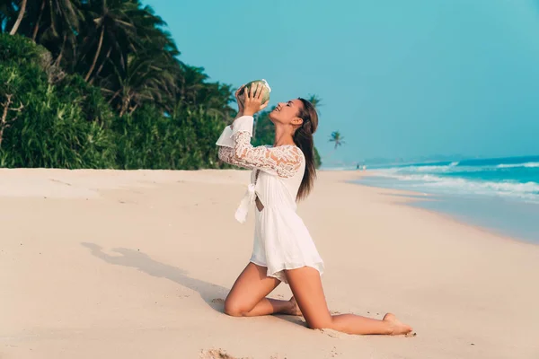 Usmívající se úhrady dívka v bílé kombinéze klečí na písečné pláži na pozadí oceánu a džungle, pití kokosové vody přímo z kokosu. — Stock fotografie