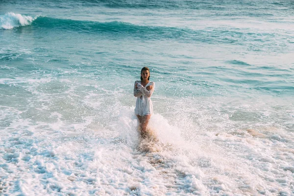 Ένα μικρό λεπτό κορίτσι σε μια όμορφη παραλία με λευκή στολή στέκεται στα γαλάζια νερά της θάλασσας, γόνατο-βαθιά. Κύματα και πιτσιλιές τύλιξε το σώμα, χέρια σταυρωμένα πάνω στο στήθος της — Φωτογραφία Αρχείου