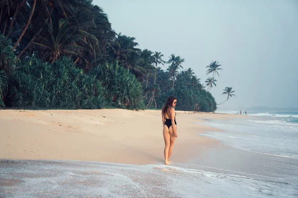 Vista desde la parte trasera de una chica chic en bikini camina por una playa desierta vacía con palmeras y grandes olas, disfrutando de la naturaleza y la belleza del océano — Foto de Stock