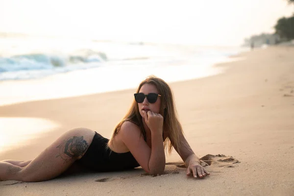 Close-up of a beautiful slender sports girl in a black bathing suit lies on the yellow soft sand by the sea, wears fashionable sunglasses. — Stock Photo, Image