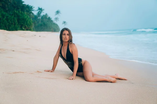 Sexy dívka v písku leží na pobřeží, se těší relaxační na prázdné bílé písčité pláži, nosí stylové černé uzavřené plavky. — Stock fotografie