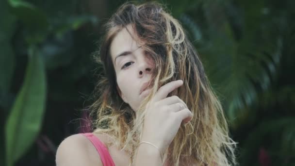 Uma imagem de close-up slow-motion de uma menina com cabelo encaracolado curto, queimado pelo sol, cobre-os com uma parte de seu rosto, senta-se no meio da vegetação tropical, fecha os olhos com prazer . — Vídeo de Stock