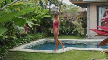 Yavaş hareket steadikam yüksek çekim kısa saçlı ince modeli havuza gidiyor. Bir kız çorap mayo bir arka plan bahçe tropik bitkilerin su gider.