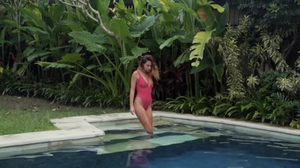 Lento-motion riprese di una ragazza snella sexy in un bellissimo costume da bagno luminoso entra in piscina, nuota e gode la sua bella vacanza presso la villa, nuota a lato della piscina ed emerge sui gomiti — Video Stock