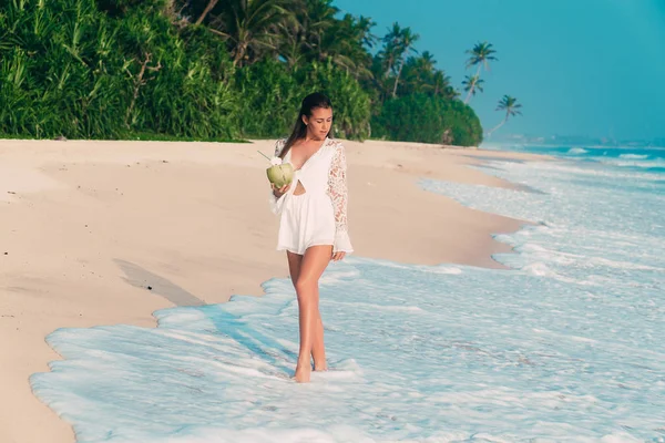 Una joven viajera europea de cabello oscuro camina por una playa de arena con un coco en la mano, una ola cubre sus piernas delgadas — Foto de Stock