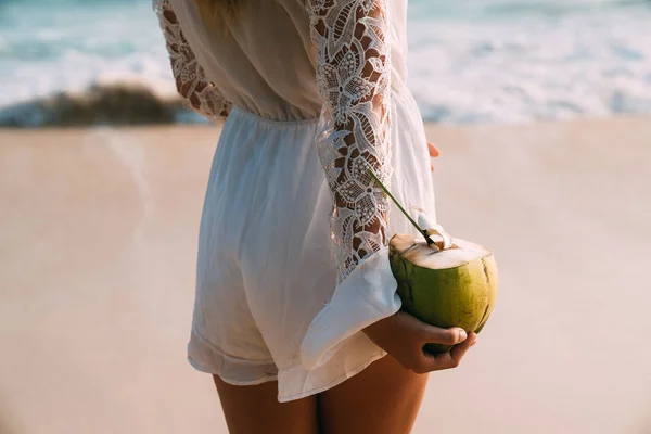 Primer plano de un marco recortado desde la parte posterior. La chica está sosteniendo un coco fresco jugoso maduro con un chasquido en la mano, bebiendo agua de coco, un culo en un general de moda . — Foto de Stock