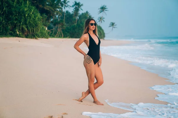 Una chica joven brillante y elegante en una playa de arena blanca con gracia va al agua, admira el surf, lleva un bikini y elegantes gafas negras — Foto de Stock