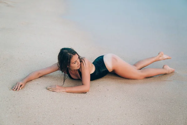 Een sexy jonge meisje met een gladde zelfs huid in een trendy zwarte zwembroek ligt op nat zand. Het concept van vrije tijd, reizen, mensen. — Stockfoto