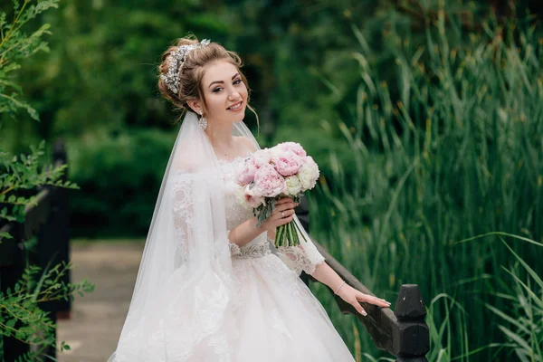 La novia está caminando a lo largo del puente sobre el río. La chica está posando en un vestido de novia y un ramo de peonías en el bosque . —  Fotos de Stock