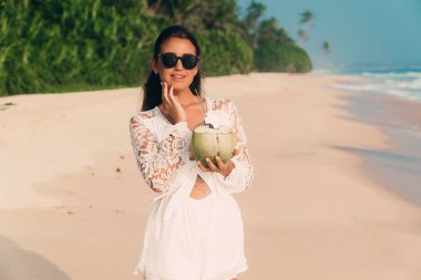 Eğlence ve seyahat kavramı. Genç bir tabaklanmış kız tesisi için geldi, şık beyaz bir elbise ve güneş gözlüğü takıyor, lezzetli bir Hindistan cevizi elinde tutan, suyu içer.