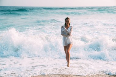 Kız yavaşça ince vücudu onunla hugs, plaj, bacaklarını su ve beyaz bir köpük dalgaların üzerinde şık plaj beyaz elbiseli güzel tabaklanmış ince parlak model duruyor