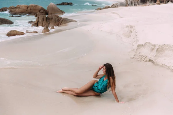 Λευκό αφρισμένα κύματα μόλις να αγγίζετε τον καιρό μαυρισμένο τα πόδια του ενός νεαρού ομορφιά που βρίσκεται στη λευκή άμμο της μια άγρια παραλία παράδεισος. Το κορίτσι διορθώνει τα μακριά μαλλιά με το χέρι και κοιτάζει το μπλε νερό. — Φωτογραφία Αρχείου