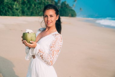 Gülümseyen güzel bir kız bir boş boş kumsala bir Hindistan cevizi bir kokteyl içer. Modeli parlar ve gülümsüyor, sahiptir tatil, deniz ve güneş