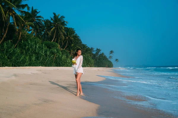 Una atractiva joven europea esbelta voló a descansar, pasea por la playa de arena, sobre el fondo de la selva y las olas del mar, bebe un coco fresco y frío . — Foto de Stock