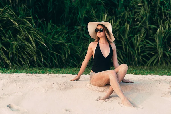 Una chica hermosa delgada joven se sienta en la arena blanca cerca de las plantas tropicales, disfruta de la relajación, baños de sol, se viste con un traje de baño caro de moda, gafas y sombrero de tendencia — Foto de Stock