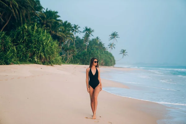 Una hermosa joven está descansando en una playa tropical, sobre un fondo de vegetación, palmeras, arena suave amarilla y mar azul.Modelo en un elegante traje de baño negro y gafas de sol —  Fotos de Stock