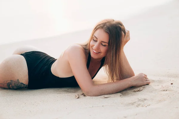 Közeli kép: egy szép boldog fiatal lány ölében nyugvó európai megjelenése. A modell fekszik a homokban, hord egy divatos kötény, és vidáman, nevet, lehunyta a szemét, öröm. — Stock Fotó