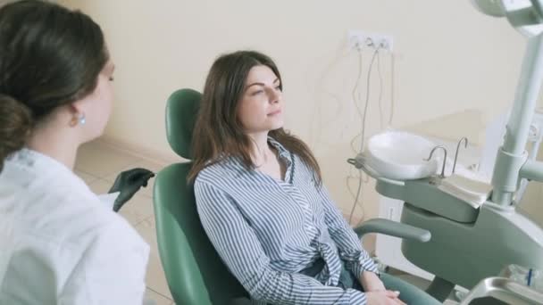 O frumoasă brunetă zâmbitoare cu păr lung în haine elegante a venit să-și trateze dinții în biroul dentiștilor. Trage încet steadikam medicul pune un șorț pe pieptul pacienților, o pregătește — Videoclip de stoc