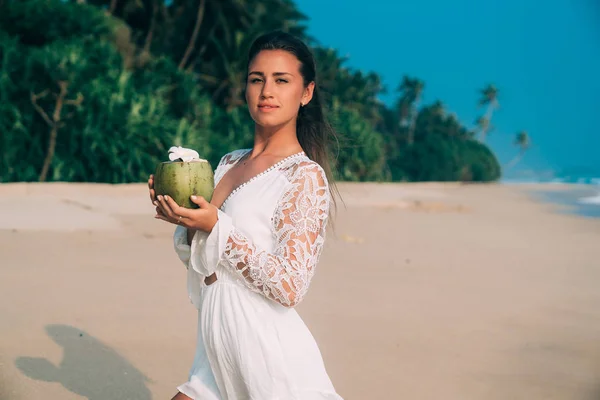 Portrét roztomilé krásné dívky v stylovém beach celkově jemně usměje a drží kokos s květinou. Vítr fouká tmavě hnědé vlasy na pláži — Stock fotografie