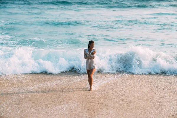 Ein schönes sexy schlankes, sonnengebräuntes Mädchen in einem weißen Strandkleid steht nass am Strand, vor dem Hintergrund von Wellen und weißem Sand, verschränkt die Arme über der Brust. das Konzept des Strandes — Stockfoto