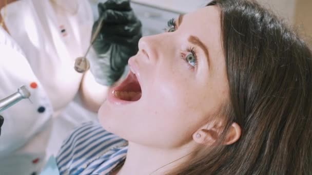 Close-up van een bijgesneden frame van een jonge mooie meisjes gezicht bij de tandarts. De patiënt ligt in de tandheelkundige stoel met een open mond, de dokter in handschoenen de tand boren met een boor en kijkt — Stockvideo