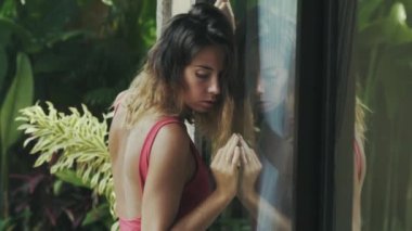 Bikinili seksi manken sıcak tropikal evinde bir cam kapı yakınında standları, ne yazık ki tepeden bakıyor ve onun bakışları yükseltir, güzel yüzü ve ince seksi vücut silüeti bardakta yansıtılır