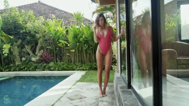 Um modelo esbelto em um traje de banho carmesim forte e elegante passeia por sua vila contra o pano de fundo de uma piscina e uma casa com uma parede de vidro, uma imagem lenta de uma garota andando em um maiô. — Vídeo de Stock