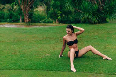 Bir kız bir ince spor tabaklanmış gövdeli bir şık siyah mayo giyer, traş çim Otelde sıcak yağmur altında oturur. Boş zaman, seyahat, yaşam kavramı.