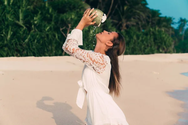 Una joven en una playa blanca y limpia bebe sexualmente jugo de un coco maduro, de pie sobre la arena vestida con un vestido blanco y aireado — Foto de Stock