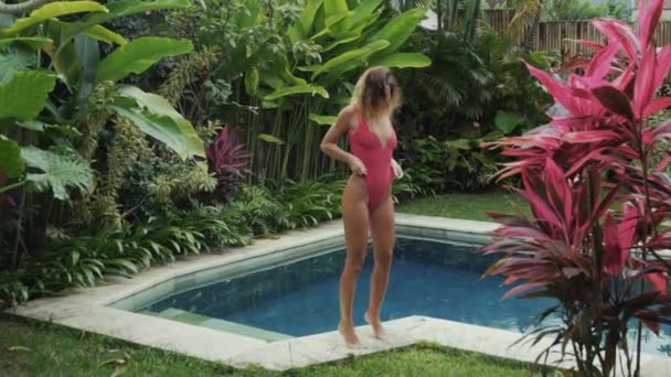 Sexy modelo bronzeado mostra seu novo maiô, vira-se nele, mostra como ele está sentado, tiro em câmera lenta, uma menina em um maiô vermelho posando perto da piscina na villa . — Vídeo de Stock